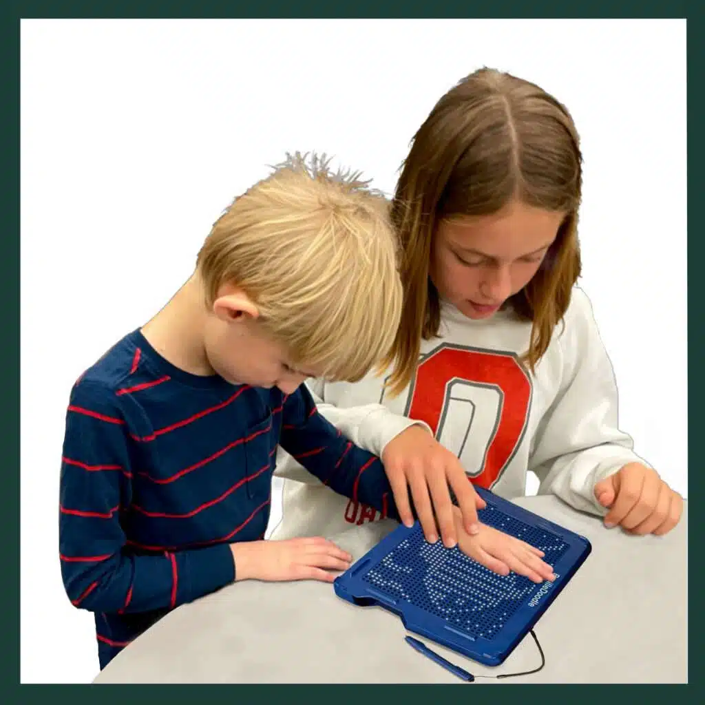 A boy with blonde hair about seven years old and wearing a blue stripe shirt, is standing with his big sister about 11 and shoulder length, with straight brown hair, in a white sweatshirt is guiding his hand on the BrailleDoodle to feel a picture.