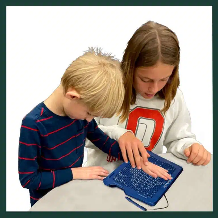 A boy with blonde hair about seven years old and wearing a blue stripe shirt, is standing with his big sister about 11 and shoulder length, with straight brown hair, in a white sweatshirt is guiding his hand on the BrailleDoodle to feel a picture.