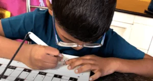 Young boy wearing a blue t-shirt and glasses in India is writing braille. He uses the magnetic stylus on the prototype of the BrailleDoodle.
