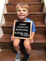 Seven-year-old gunner is sitting on some steps holding a plaque that says second grade and smiling widely