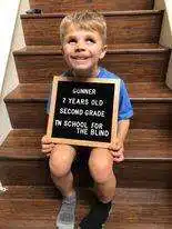 Seven-year-old gunner is sitting on some steps holding a plaque that says second grade and smiling widely