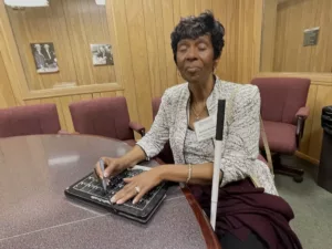 Board of director at NFB-An African-American woman with short dark hair wearing an elegant black and white jacket and dress is forming Braille and the BrailleDoodle.