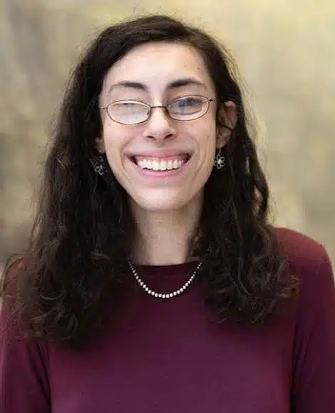A woman with long hair, wearing glasses, a maroon sweater, a pearl necklace is smiling widely into the camara