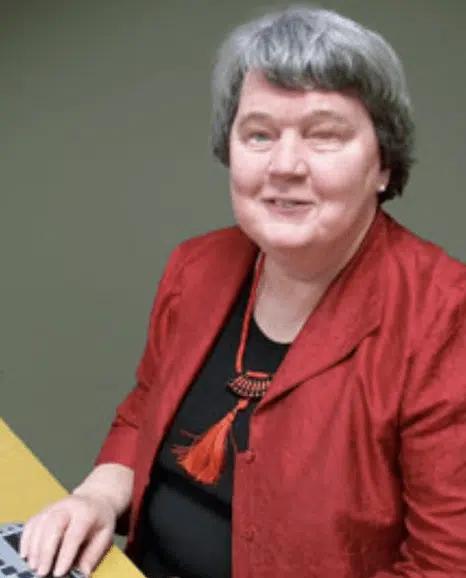 A caucation woman with short gray hair , wearing a red jacket over a black top with a red tassel necklace, smiles while seated at a table.