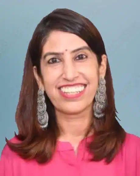A woman of Indian dissent is smiling broadly at the camera. She is wearing long, elaborate earrings, and a fuchsia blouse. She has long straight dark hair that falls below her shoulders.