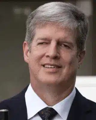 Middle-aged man smiling with grey hair, in a dark suit is holding a white cane